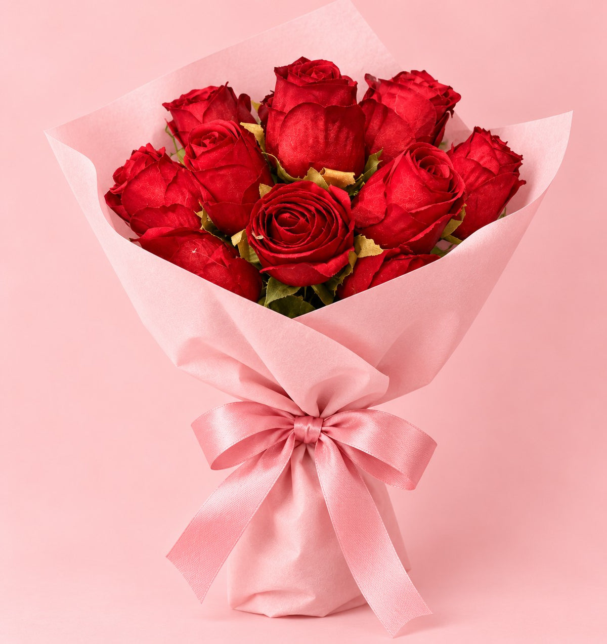 A bouquet of 12 red roses wrapped in light pink tissue paper and tied with a matching pink satin bow, set against a solid pink background.
