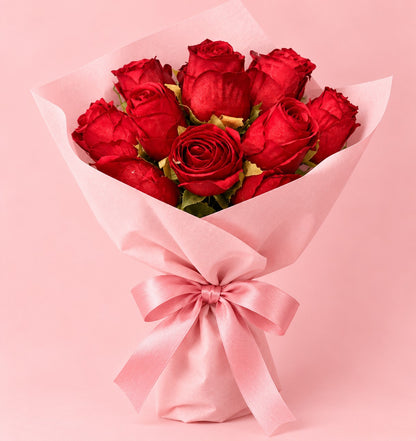 A bouquet of 12 red roses wrapped in light pink tissue paper and tied with a matching pink satin bow, set against a solid pink background.
