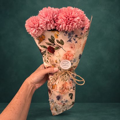 A hand holding a bouquet of pink carnations wrapped in floral-patterned paper with a round &