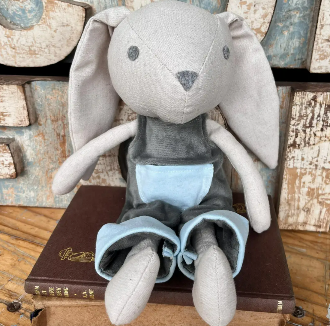 Australian-made cotton plush bunny with long floppy ears, in grey dungarees with a pale blue pocket, sitting on a stack of vintage books