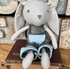 Australian-made cotton plush bunny with long floppy ears, in grey dungarees with a pale blue pocket, sitting on a stack of vintage books