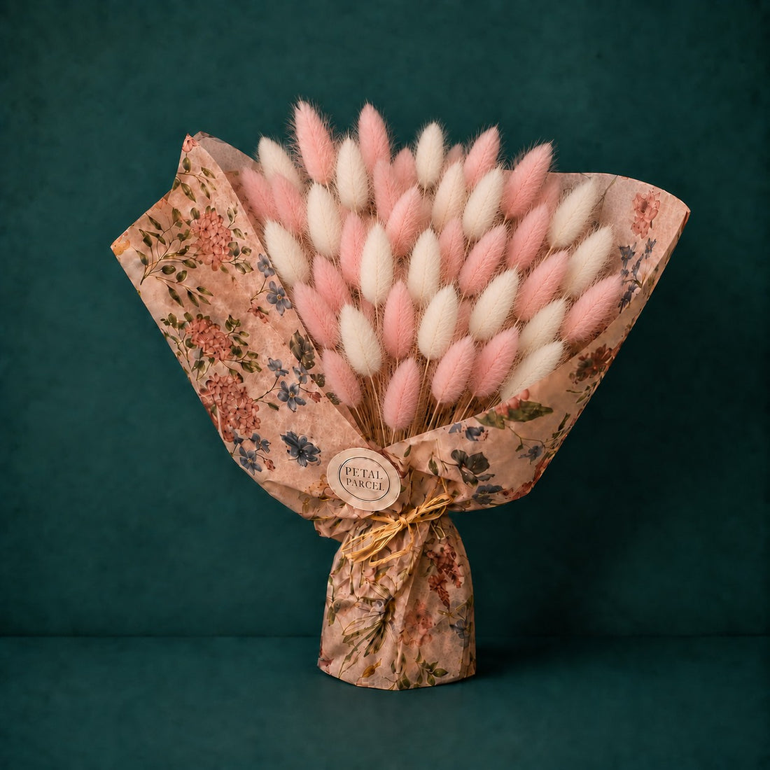 A bouquet of dried bunny tail grass stems wrapped in floral paper, with pink and white fluffy stalks fanned out against a dark teal background. A round label at the center reads &