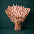 A bouquet of dried bunny tail grass stems wrapped in floral paper, with pink and white fluffy stalks fanned out against a dark teal background. A round label at the center reads &