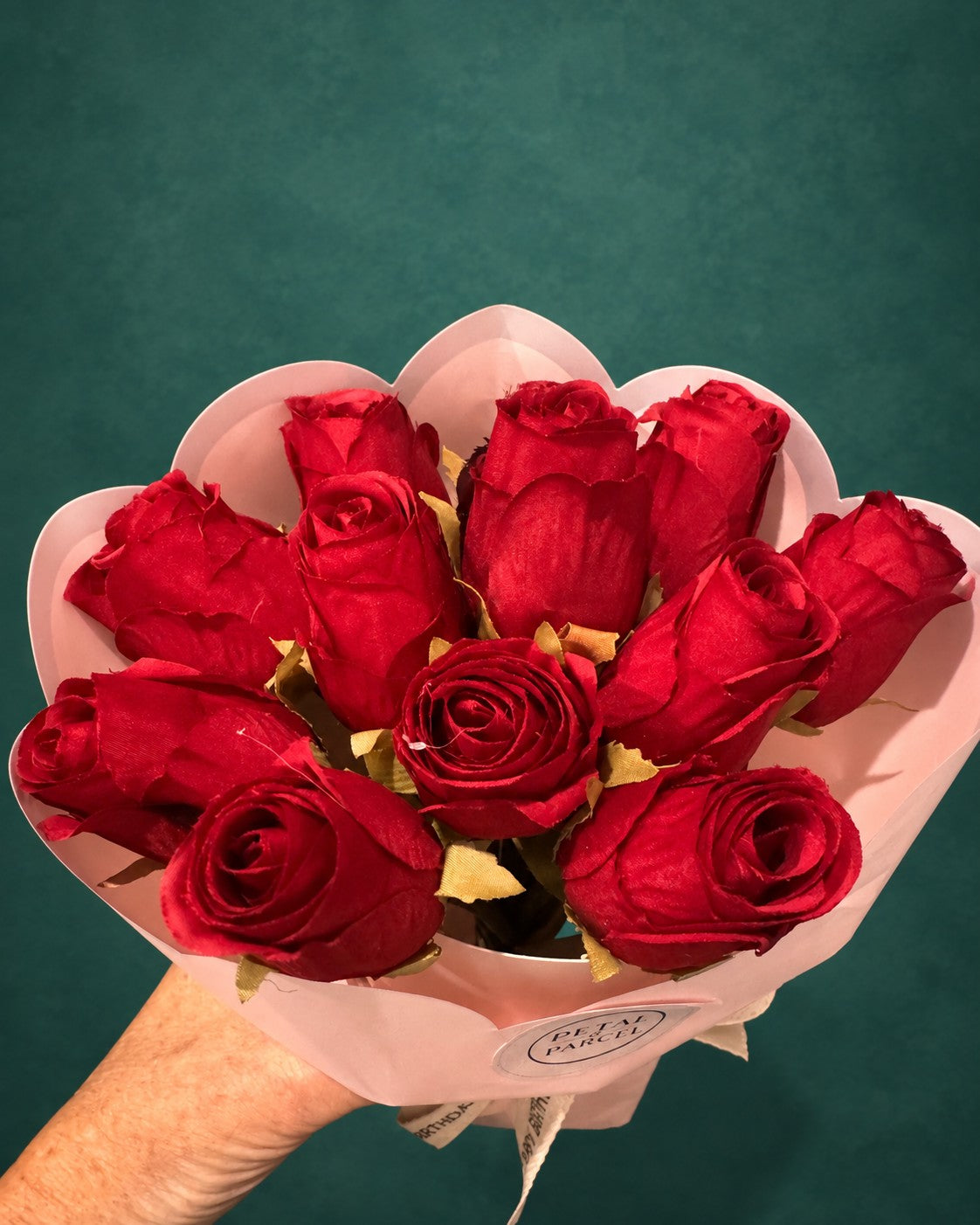 A bouquet of ten red roses wrapped in light pink paper, held by a hand against a dark teal background.