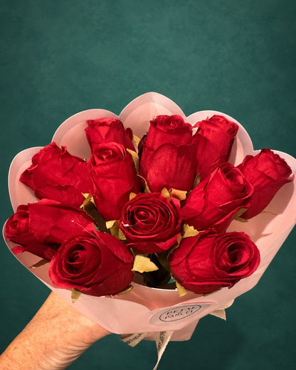 A bouquet of ten red roses wrapped in light pink paper, held by a hand against a dark teal background.