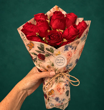 A hand holding a bouquet of red roses wrapped in decorative floral tissue paper with a round &
