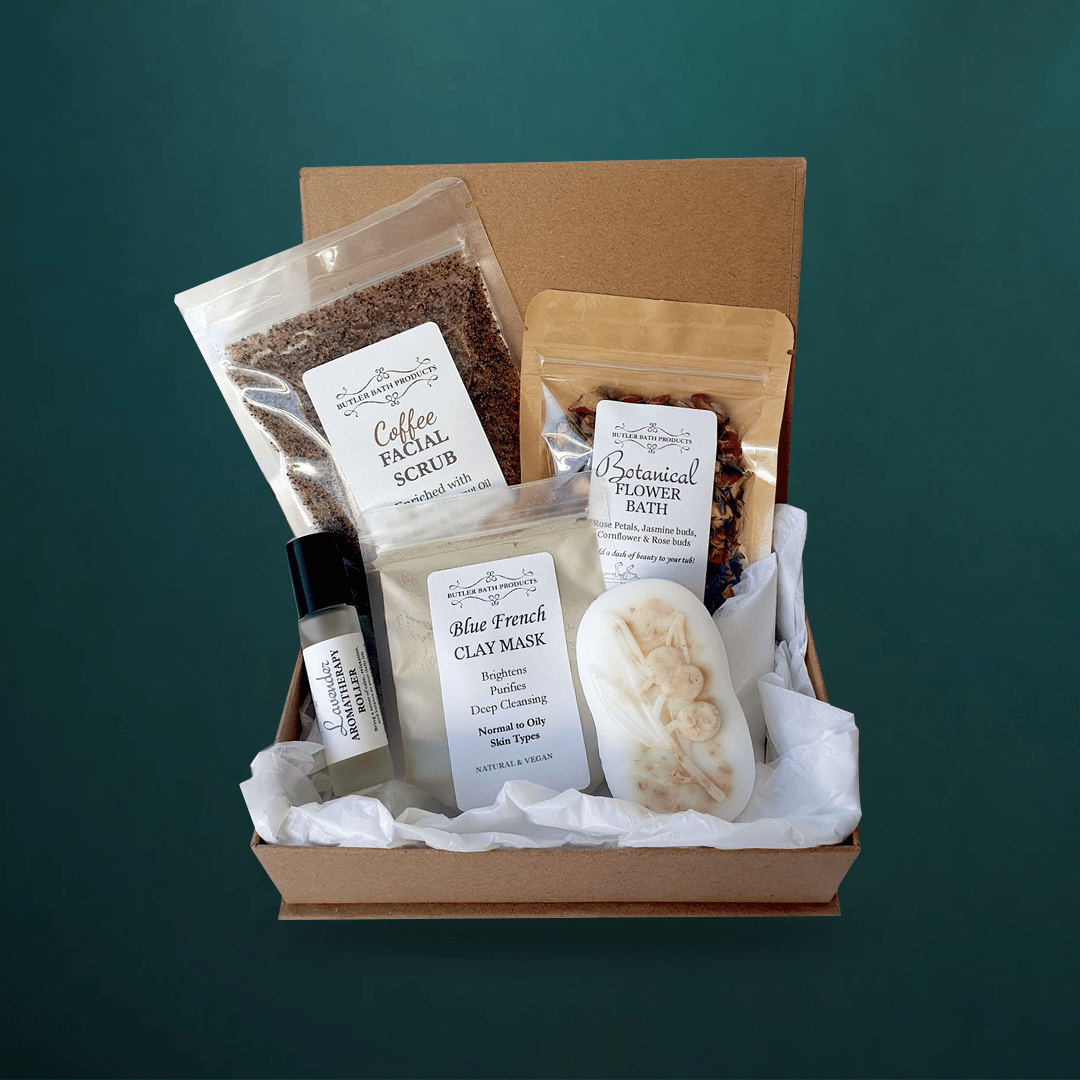 Five-piece Butler Bath pamper pack with coffee facial scrub, blue French clay mask, oatmeal soap, lavender roller and botanical flower bath in a kraft gift box, on dark teal